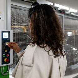 Eine Person bedient das Steuerungspanel eines halbautomatischen Parksystems, um das geparkte Fahrzeug abzurufen. Im Hintergrund sind die verschiebbaren Stellplätze und ein Fahrzeug innerhalb der Parkanlage sichtbar.