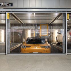 L'immagine mostra un'auto sportiva gialla in un sistema di parcheggio all'interno di un moderno parcheggio sotterraneo.